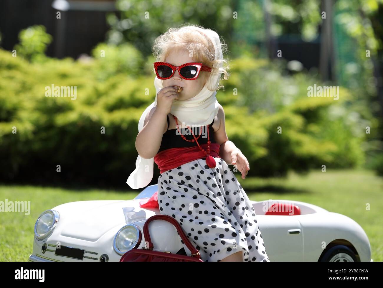 Porträt eines hübschen kleinen blonden Mädchens mit einem weißen Taschentuch, das sich an einem weißen Spielzeugauto im Garten lehnt Stockfoto