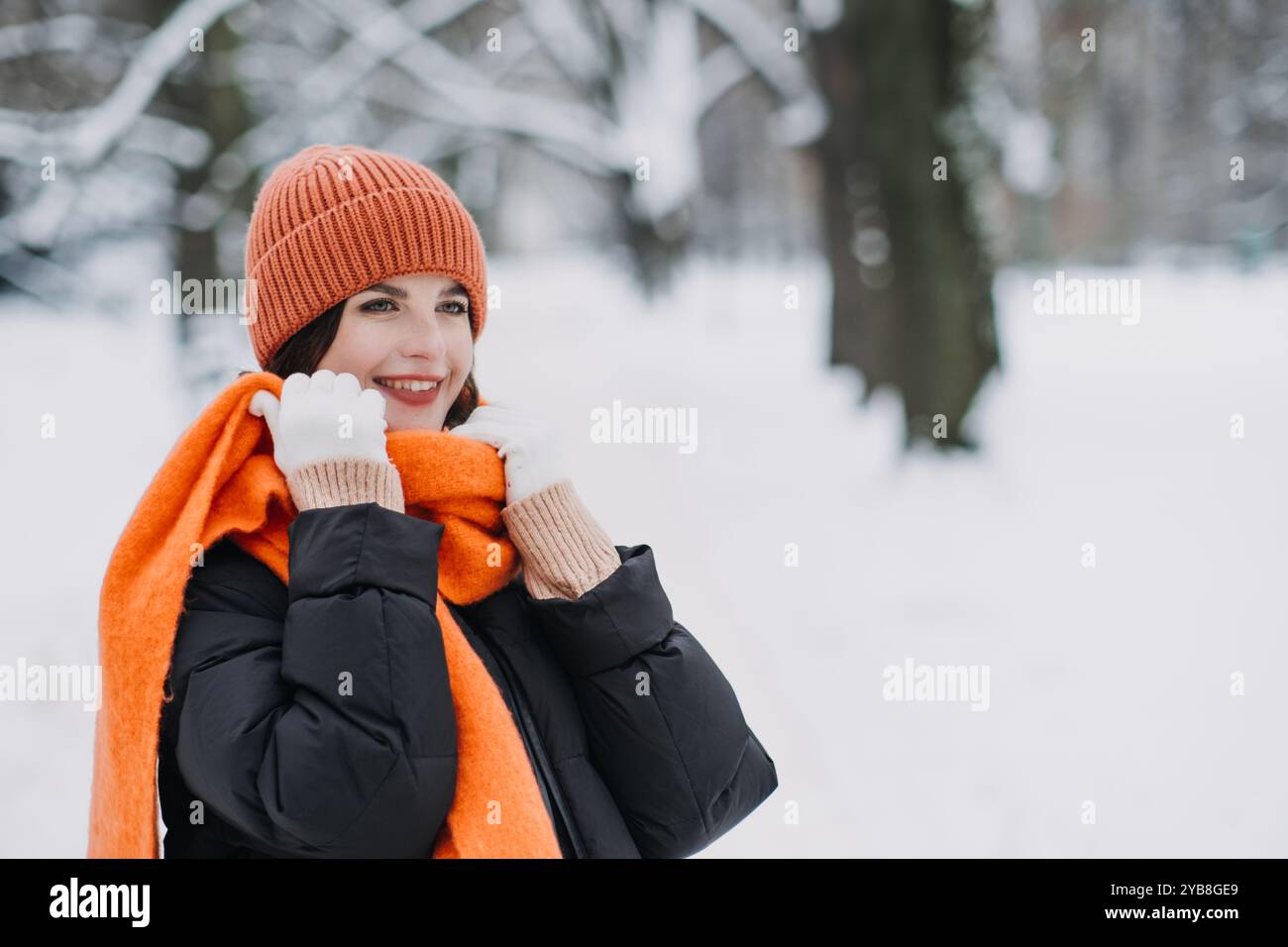 Frau mit orangefarbenem Winterhut und Schal lächelt im schneebedeckten Park. Warme saisonale Mode, winterliche Trends, gemütliche Outdoor-Bekleidung. Farbtherapie in Stockfoto