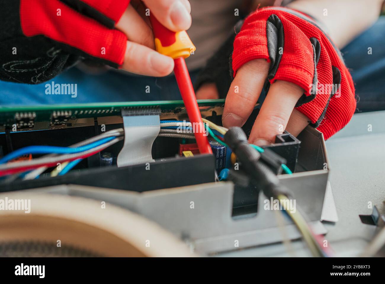 Elektriker in Handschuhen arbeitet akribisch auf einer Leiterplatte und verbindet Drähte mit einem Schraubendreher. Die Nahaufnahme zeigt geschickte Hände, die ein kaputtes Gerät präzise fixieren Stockfoto