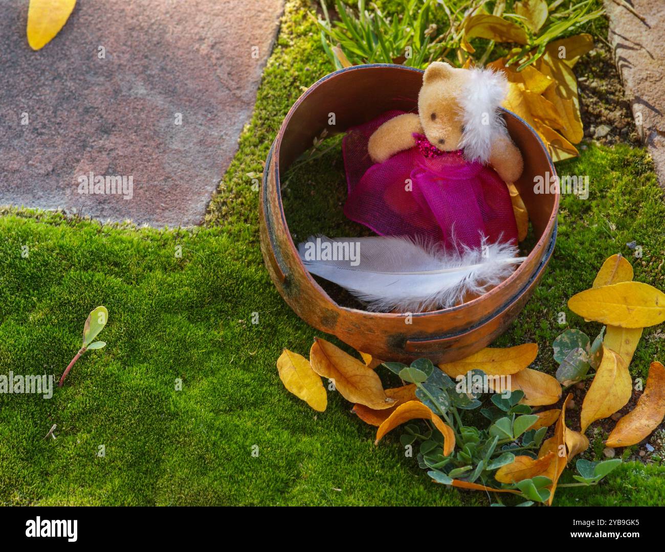 Die Miniatur-Ausstellung auf grünem Moosteppich zeigt einen flauschigen Teddybären mit weißen Federn, umgeben von Blättern. Schieferfertiger machen Winkel auf dem Gartenweg. Stockfoto