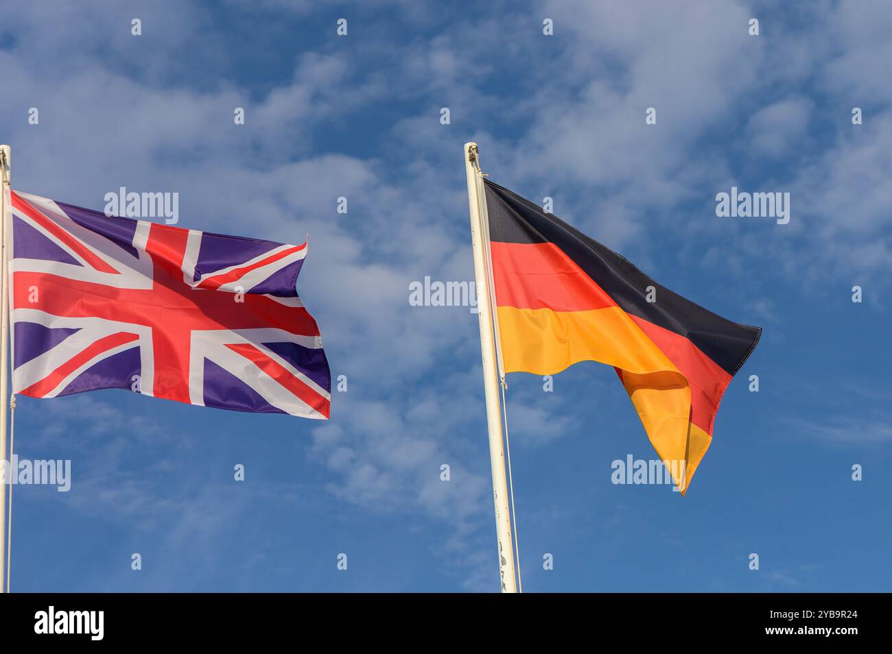 Zwei leuchtende Flaggen, die das Vereinigte Königreich und Deutschland repräsentieren, winken sanft vor einem Hintergrund aus flauschigen Wolken und hellblauem Himmel und symbolisieren Freunde Stockfoto