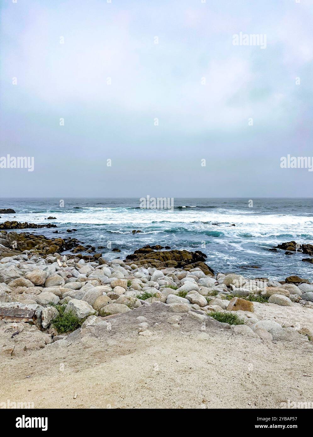 Bewölkter Tag am Sandstrand Pebble Beach in Kalifornien mit blauen Wellen, die in braune Felsen stürzen und im Herbst Meeresschaum erzeugen. - Smartphone-aufgenommenes Stockfoto
