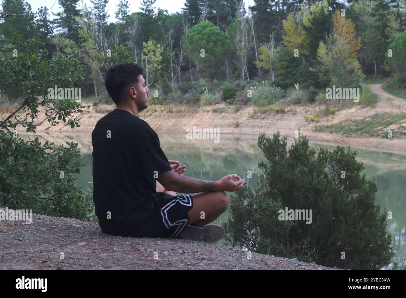 Ein junger Mann sitzt friedlich an einem ruhigen See und meditiert in einem ruhigen, reflektierenden Zustand. Die ruhige Szene fängt die Harmonie zwischen Körper und Natur ein Stockfoto