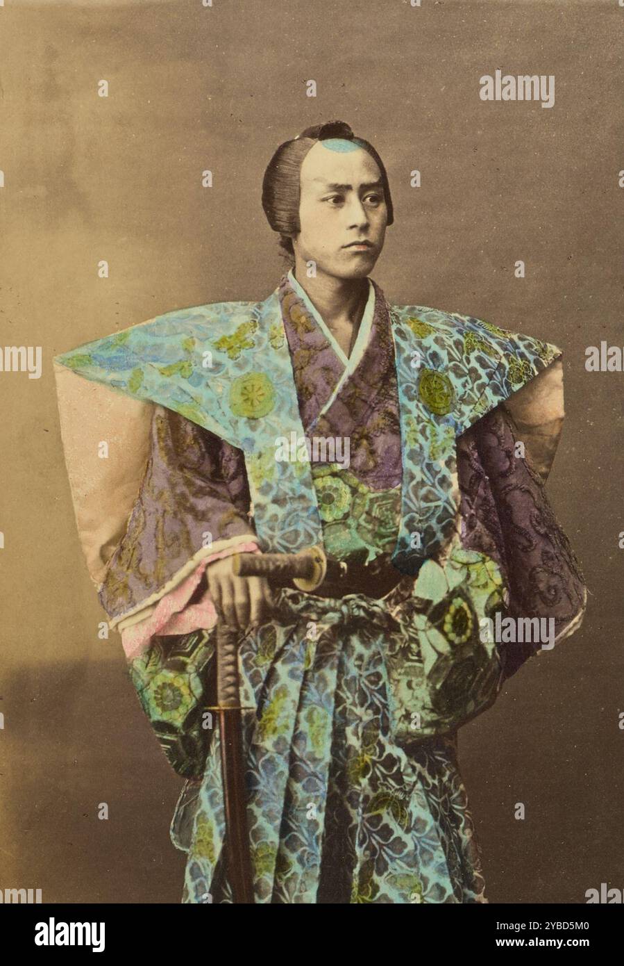 Japanischer Krieger, 1868-1880. Studio-Porträt eines Mannes in traditioneller Samurai-Kleidung. Er trägt ein aufwändig gemustertes Kamishimo und trägt zwei Schwerter. Eines der Schwerter ist in seinem Gürtel verstaut und das andere hält er aufrecht mit der Hand auf dem Pommel. Stockfoto