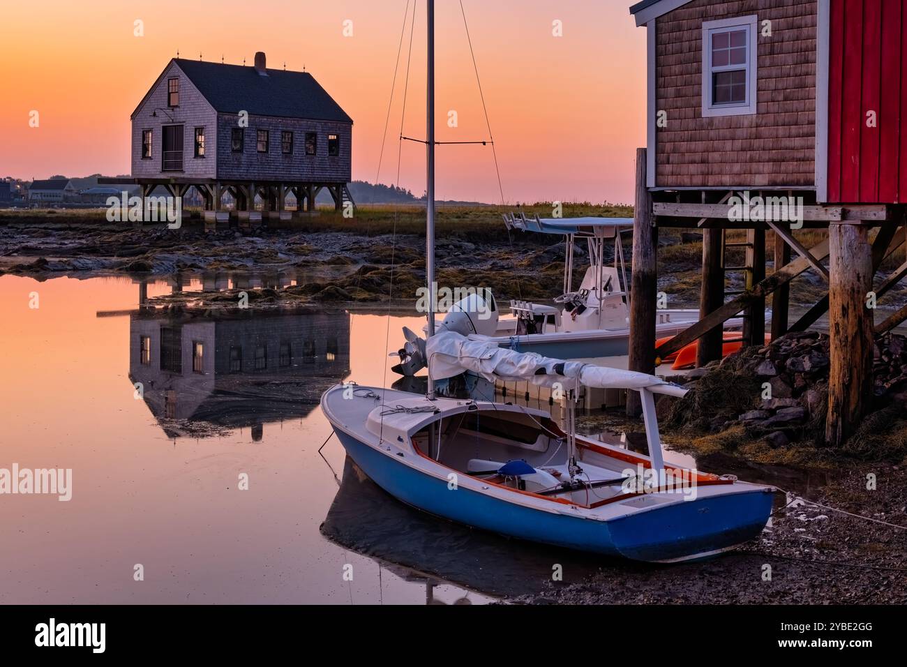 Die ruhige Küstenlandschaft in der Abenddämmerung zeigt Boote, die an Holzgebäuden in der Nähe des ruhigen Wassers angedockt sind und die lebendigen Farben des Sonnenaufgangs reflektieren. Stelzenkonstruktion Stockfoto