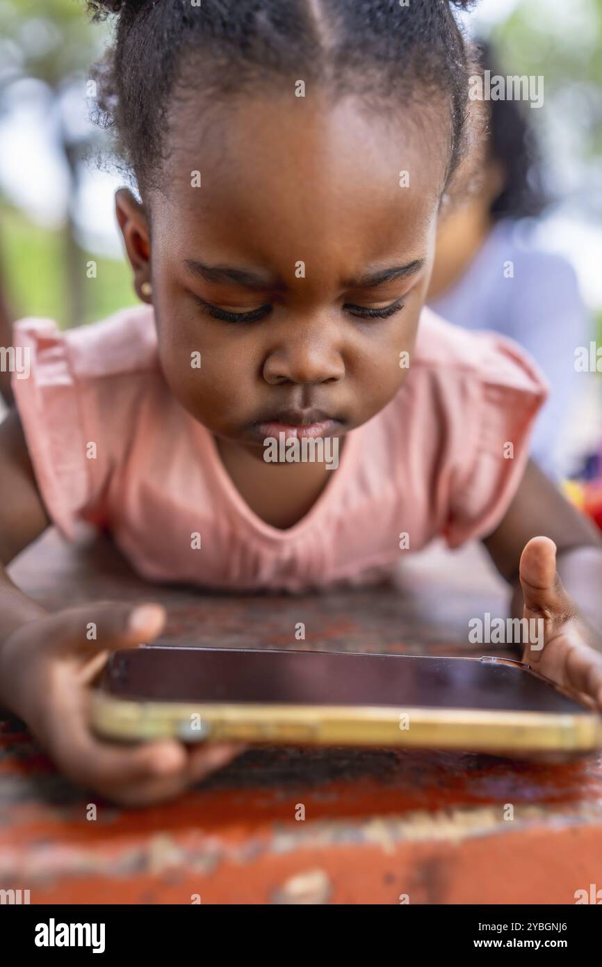 Vertikales Nahaufnahme eines afrikanischen Mädchens, das süchtig nach dem Bildschirm eines Mobiltelefons ist, das im Park neben der Familie liegt Stockfoto