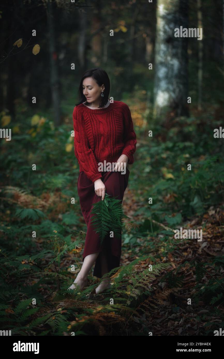 Die Frau wandert anmutig durch einen lebendigen Wald, gekleidet in einem gemütlichen roten Pullover und einem kastanienbraunen Rock, wobei sie sanft einen grünen Farn inmitten des bunten Autors umklammert Stockfoto