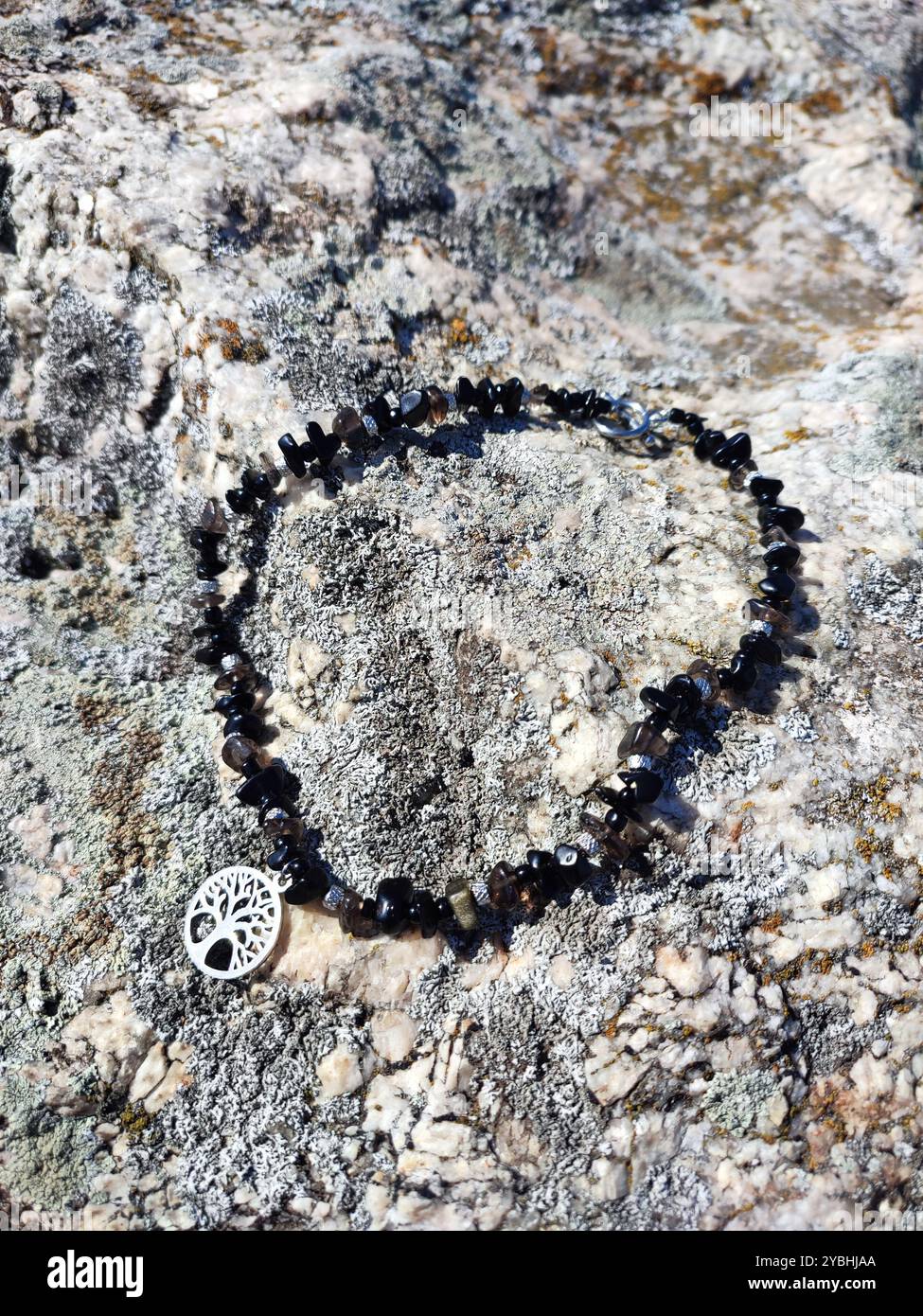 Schwarzer Obsidian Choker mit Baum des Lebens Anhänger an grauer Granitfelsenoberfläche | Chunky Halskette aus Naturstein mit Baum des Lebens Charme - Smartphone-aufgenommenes Stockfoto