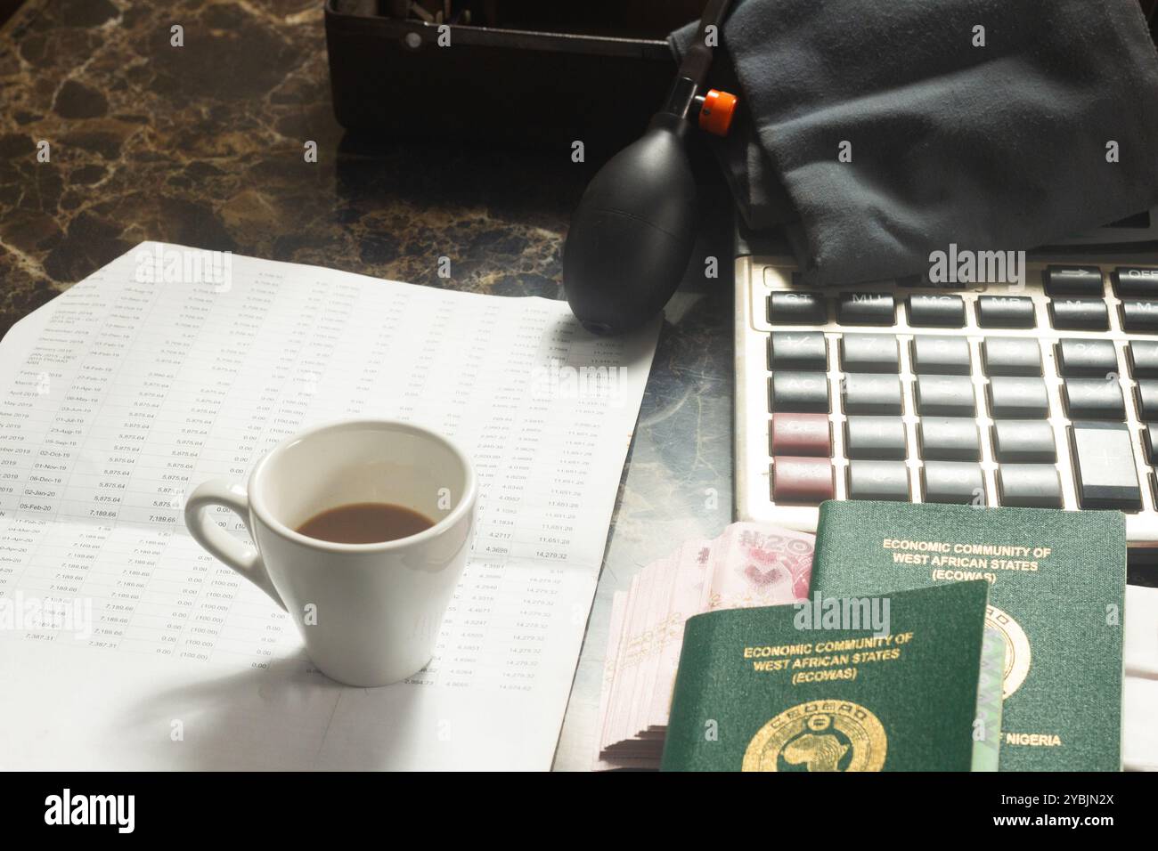 Internationaler Reisepass, Naira, Brieftasche, Kaffee, Taschenrechner, Blutdruckmanschette, Marmortisch symbolisiert Reisen, Währung, Finanzen, Geld, Gesundheit Stockfoto