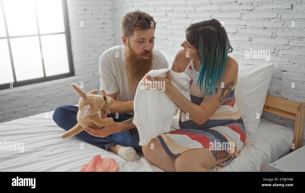 Schwangere Frau und Mann sitzen zusammen auf einem Bett in einem Schlafzimmer und teilen einen zärtlichen Moment, während sie sich auf ihr Baby vorbereiten, indem sie einen Einteiler und einen stu betrachten Stockfoto