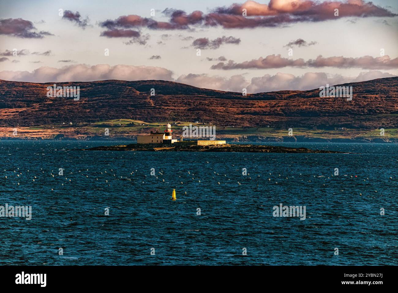 Ein Leuchtturm steht auf einer kleinen Insel, umgeben von einem blauen Meer. Der Hintergrund zeigt sanfte Hügel und einen Himmel voller leichter Wolken. Stockfoto