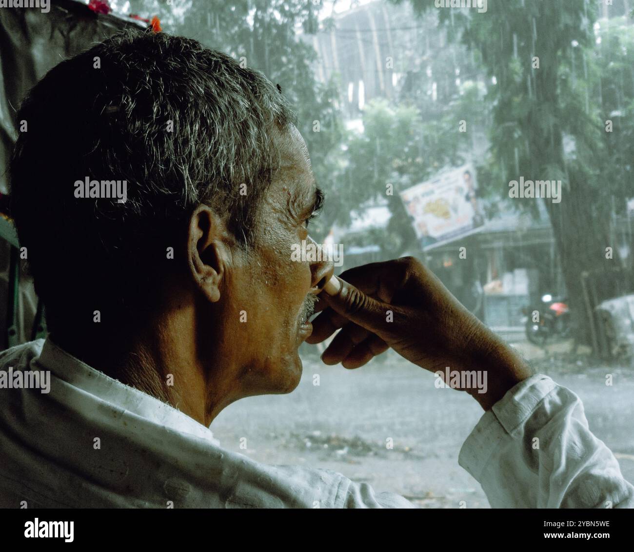 Ein alter Mann, der auf einem Stuhl sitzt und in Gedanken verloren ist, während er draußen in Bangladesch den Regen beobachtete. Ein Straßenfoto von einem einsamen Mann. Stockfoto