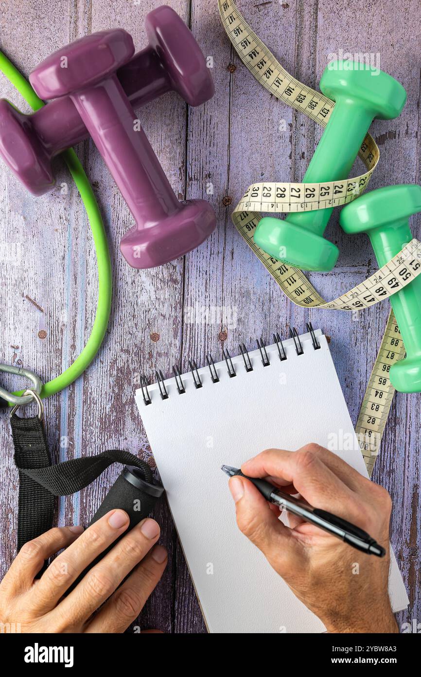 Zwei Hände und ein Notizblock, Kurzhanteln, Gummibänder und ein Maßband, Selbstverbesserungskonzept und Vorsätze für das neue Jahr Stockfoto