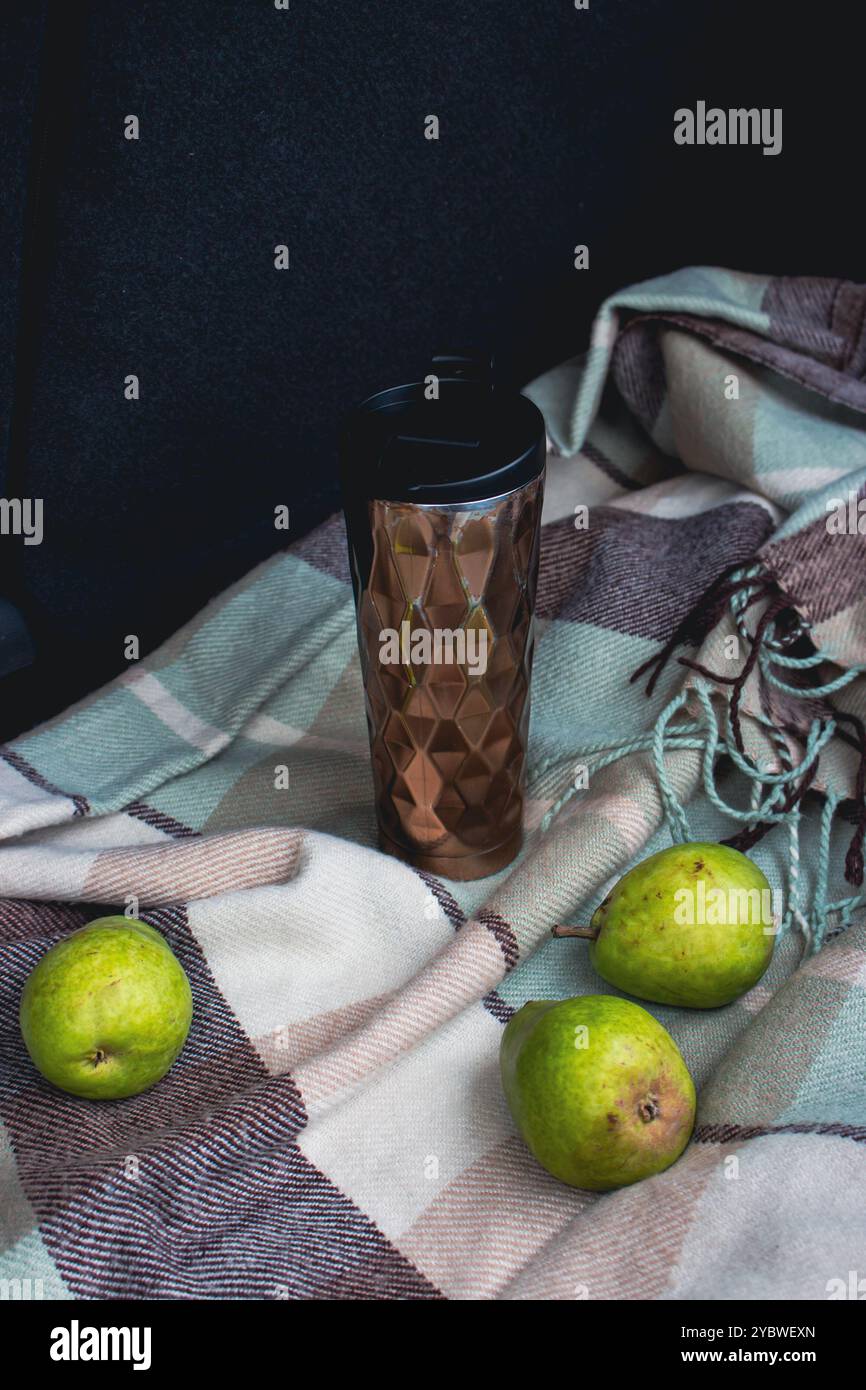 Drei Birnenfrüchte und Reisebecher auf karierter Decke im offenen Kofferraum. Stillleben im Herbst. Picknick auf dem Roadtrip. Lifestyle-Momente auf Reisen. Stockfoto