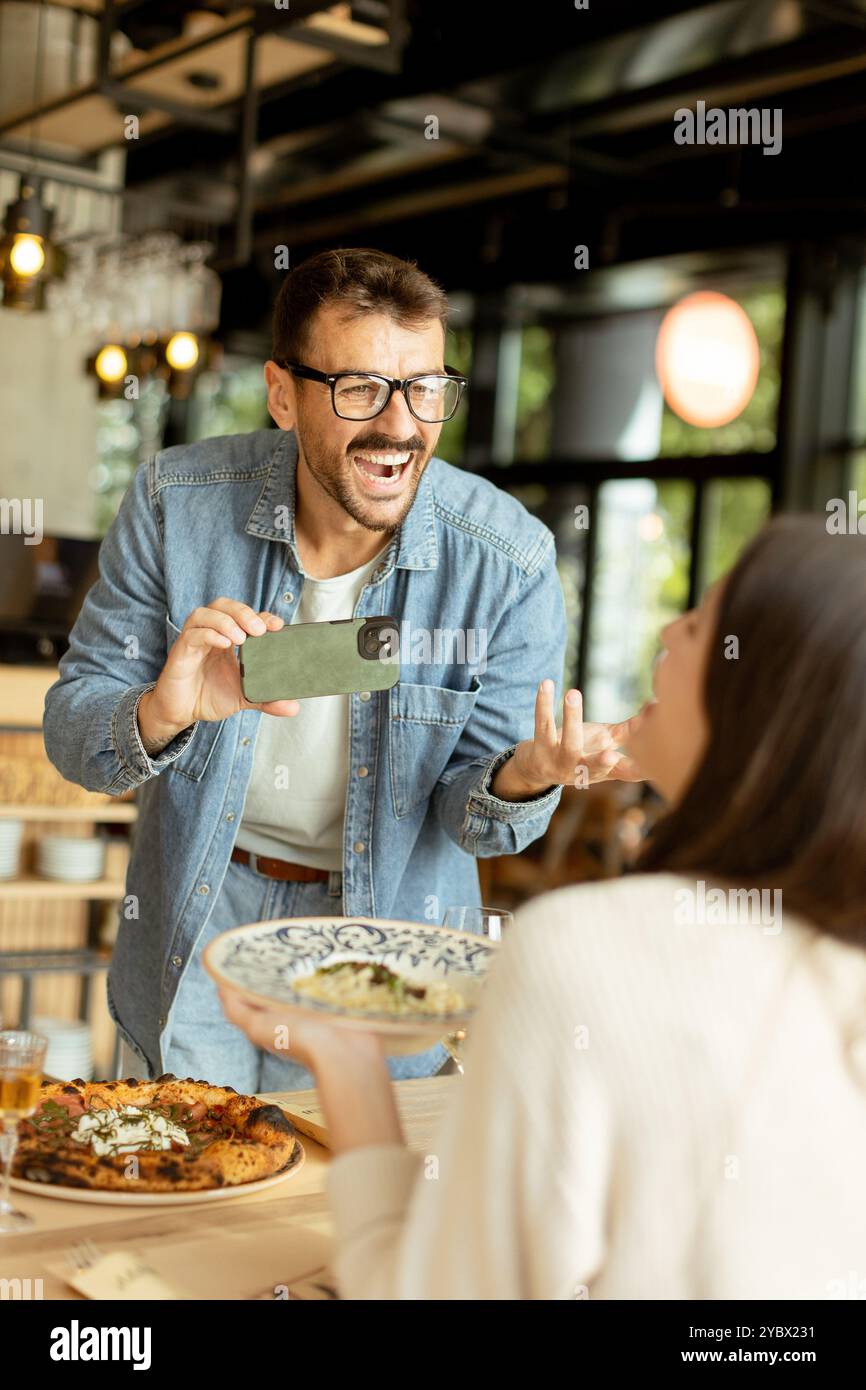 Ein fröhliches Paar sitzt an einem charmanten Café-Tisch, lächelt, während man den Moment auf einem Smartphone festnimmt, umgeben von köstlichem Essen und sonnigem Ambiente Stockfoto