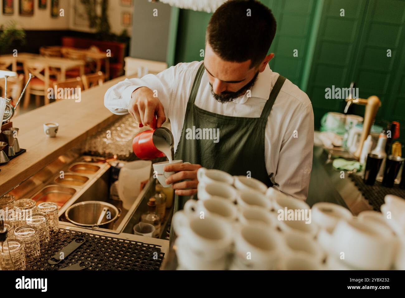 In einem warmen und einladenden Café gießt ein engagierter Barista fachmännisch Milch in eine Kaffeetasse und sorgt so für ein angenehmes Erlebnis für Gäste. Stockfoto