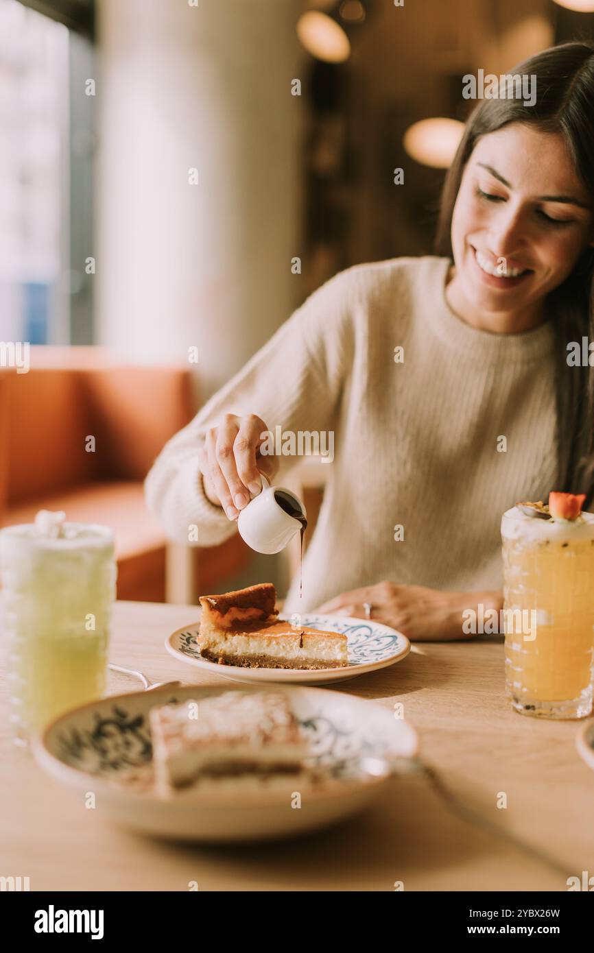 In einem charmanten Café spritzt eine lächelnde Frau gekonnt Sirup über eine dekadente Scheibe Käsekuchen und genießt eine herrliche Leckerei mit Wärme und Stockfoto