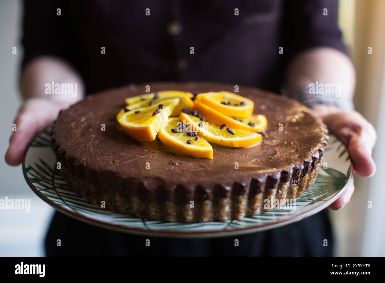 Frauenhände halten eine reichhaltige, hausgemachte Schokoladentorte mit frischen Orangenscheiben und Kakaospitzen für einen Hauch von Geschmack. Stockfoto