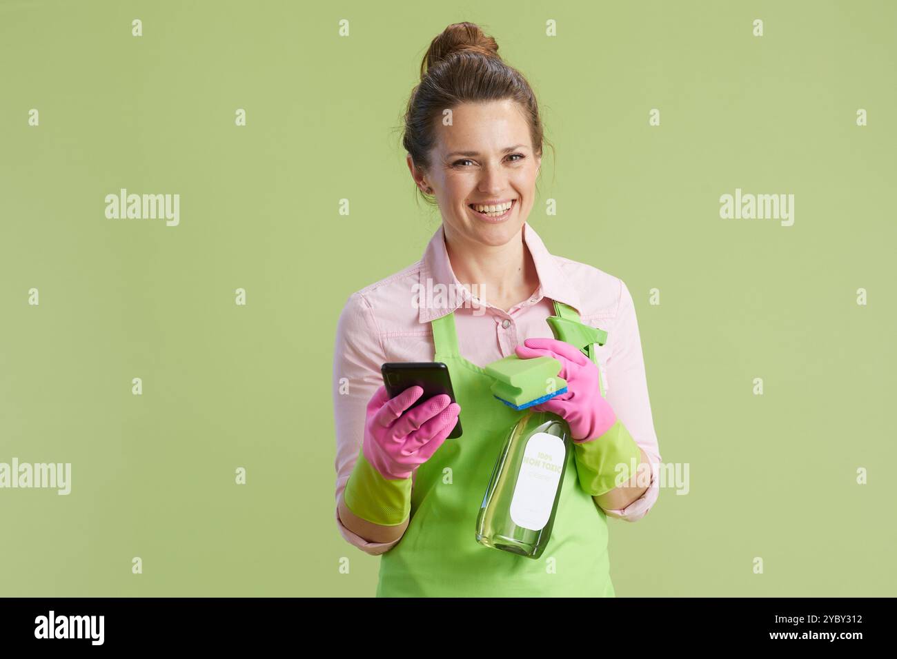 Frühjahrsputz. Glückliche moderne Frau in grüner Schürze und Gummihandschuhen auf grünem Hintergrund mit Flasche organischem Reinigungsprodukt und Smartphone. Stockfoto