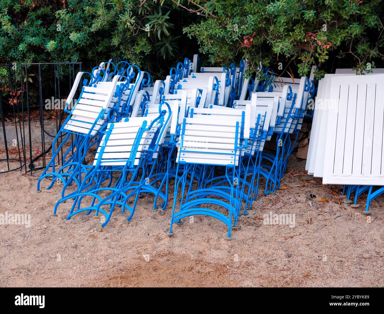 Viele weiß-blau gefaltete Holzstühle mit metallischen Beinen und Tischen, die auf Sand in der Nähe grüner Büsche gestapelt sind Stockfoto