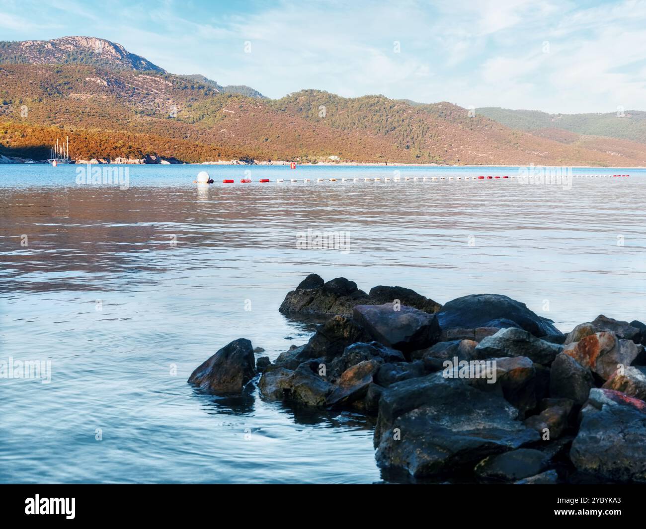 Ruhiges Meer mit Felsen im Vordergrund und Bojen, die den Badebereich mit bewaldeten Bergen im Hintergrund markieren Stockfoto