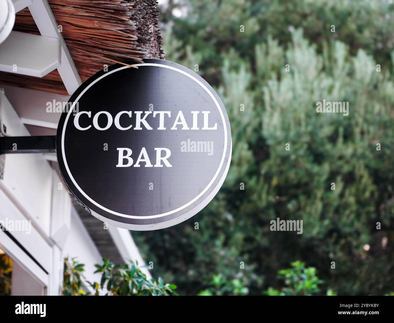 Rundes schwarz-weißes Schild für die Cocktailbar, das vor einem Gebäude mit Strohdach hängt Stockfoto