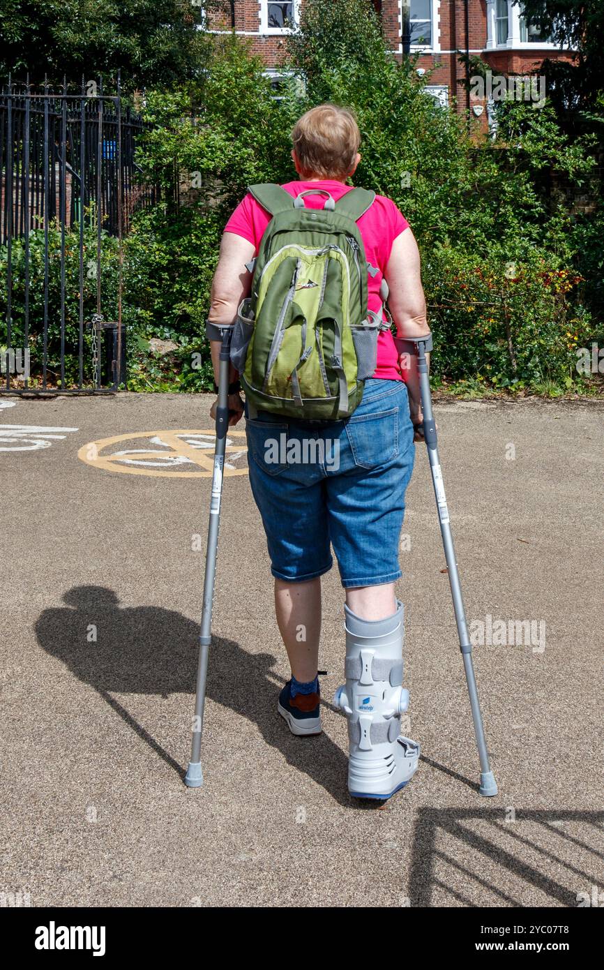 Eine Frau mit gebrochenem Bein in einem Schutzstiefel auf Krücken in einem Park, London, Großbritannien Stockfoto