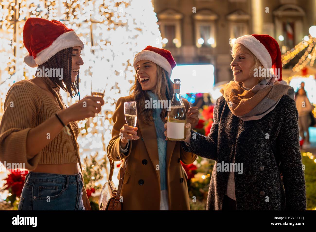 Drei verschiedene Frauen mit Weihnachtsmützen genießen festliche Getränke auf dem Weihnachtsmarkt. Freunde verschiedener Ethnien und Alters teilen sich Champagner und lachen, togeth Stockfoto