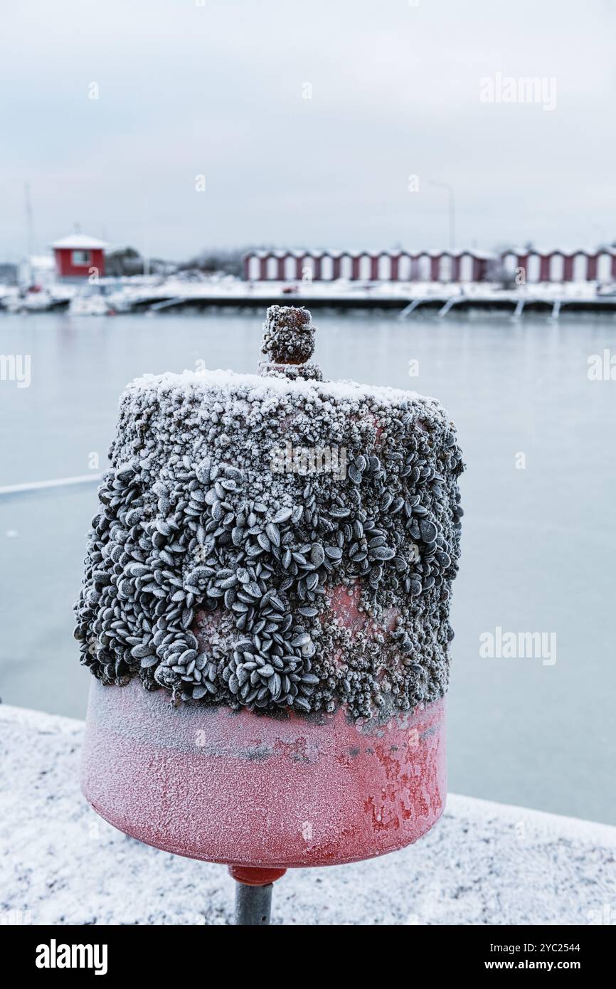 Am Wasserrand steht eine Boje, beschichtet mit einer Schicht aus Seebrücken und Schnee, mit einem gefrorenen Hafen und roten Bootshäusern, die während einer im Hintergrund sichtbar sind Stockfoto