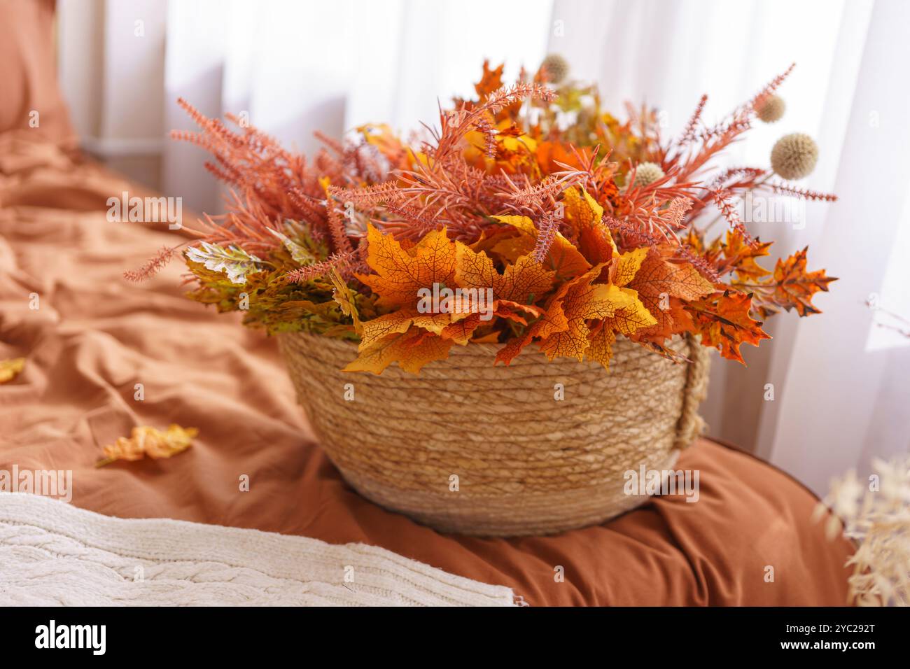 Schöne Anordnung von orange herbstfarbenen Blättern und Blumen wird in gewebtem Korb gezeigt. Der Korb liegt auf einem Bett in warmen Farbtönen und ergänzt das gemütliche Ambiente Stockfoto