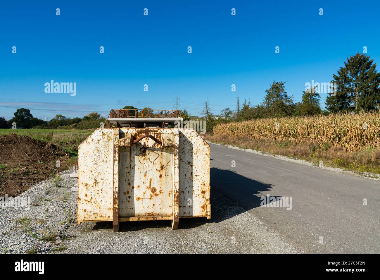 Ein rostiger Behälter befindet sich am Rande einer Schotterstraße neben einem Maisfeld unter einem klaren blauen Himmel, der den Lauf der Zeit und Vernachlässigung im Kuu widerspiegelt Stockfoto