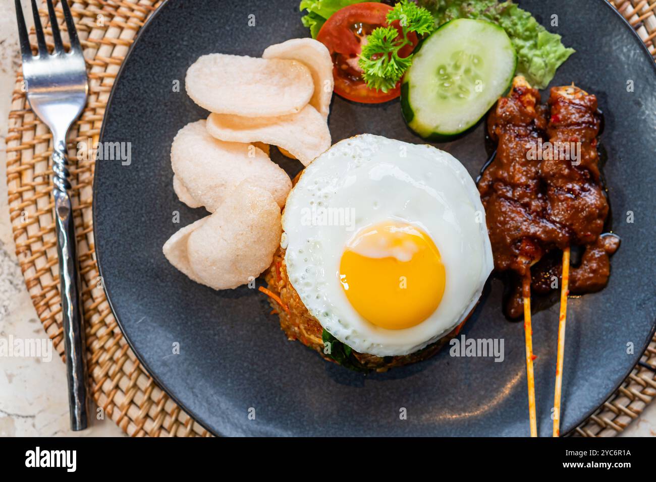 Ein Teller mit gebratenem Reis, garniert mit einem sonnigen Ei, serviert mit Gurkenscheiben, Salat und Garnelencrackern, dazu Satay-Spieße mit Pean Stockfoto