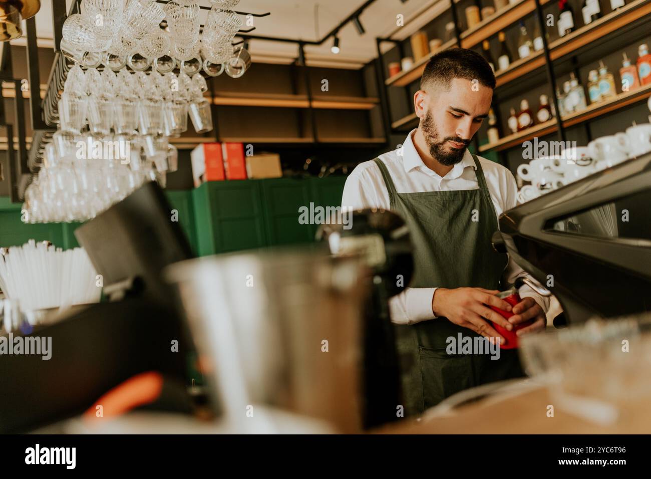In einem schicken Café konzentriert sich ein erfahrener Barista auf die Herstellung köstlicher Getränke, umgeben von ordentlich angeordneten Gläsern und lebendiger Einrichtung, die eine warme Atmosphäre schaffen Stockfoto