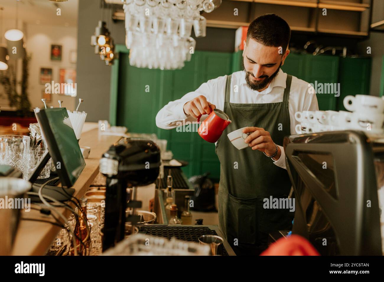 In einem warmen und einladenden Café gießt ein engagierter Barista fachmännisch Milch in eine Kaffeetasse und sorgt so für ein angenehmes Erlebnis für Gäste. Stockfoto