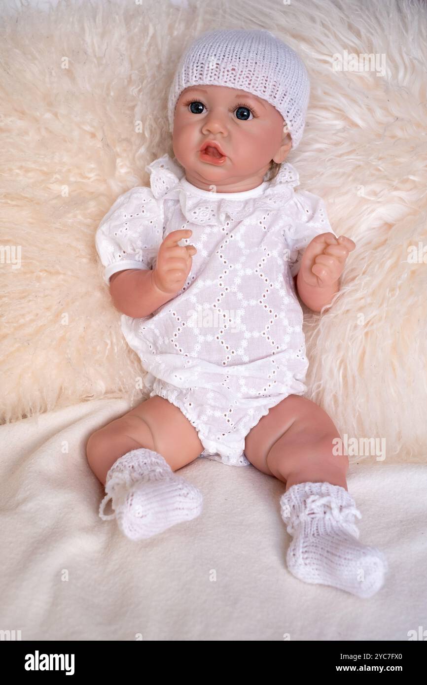 Eine lebensechte Babypuppe sitzt leise auf einer weichen, strukturierten Decke und trägt ein weißes Outfit, ergänzt durch eine Strickmütze und passende Schühchen für eine gemütliche Tappe Stockfoto