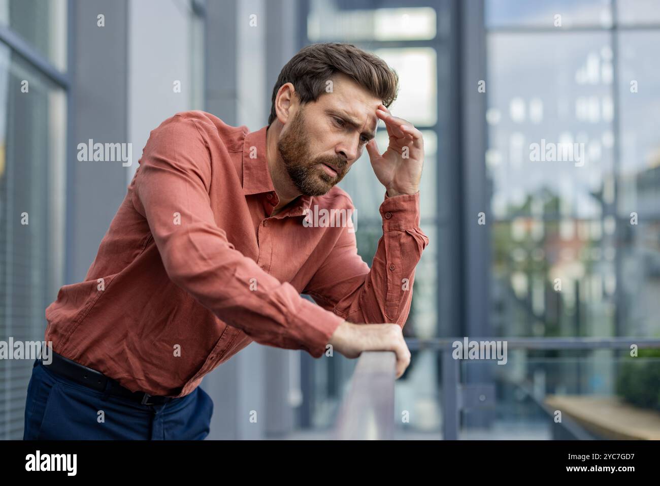 Mann mit Stress und Sorge, gestützt auf Balkongeländer in Geschäftskleidung. Das berufliche Umfeld deutet auf arbeitsbezogene Herausforderungen und Kontempla hin Stockfoto