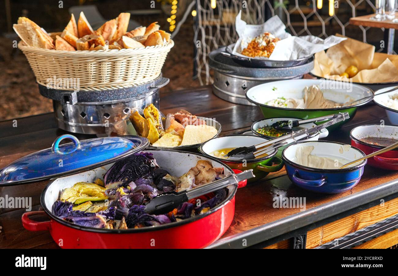 Ein Buffet-Tisch im Freien mit einem Korb mit Brot, verschiedenen Dips und farbenfrohem gegrilltem Gemüse in Schüsseln. Stockfoto