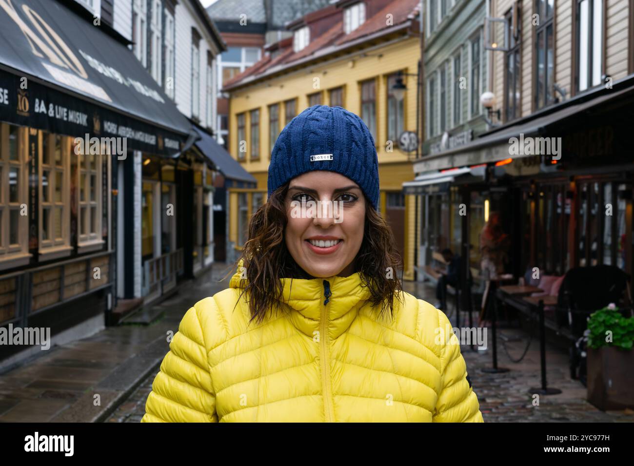 Eine junge Brünette, etwa 30 Jahre alt, trägt eine gelbe Pufferjacke und einen blauen Strickmütze, lächelt direkt in die Kamera, während sie auf einem rai steht Stockfoto