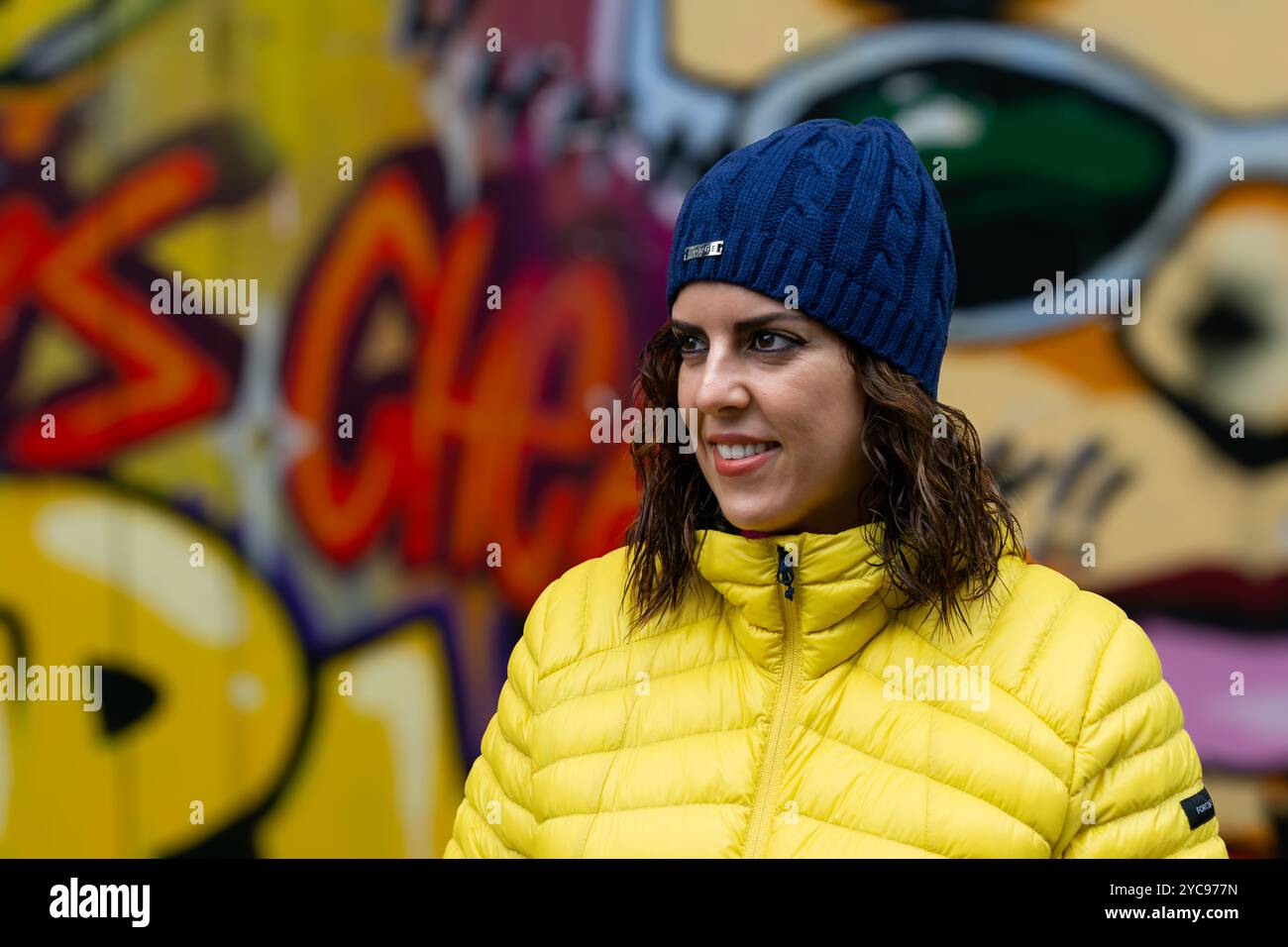 Eine junge Brünette, etwa 30 Jahre alt, in einem gelben Anorak und einer blauen Wollmütze gekleidet, lächelt zur Seite, während sie mit einem graffi steht Stockfoto