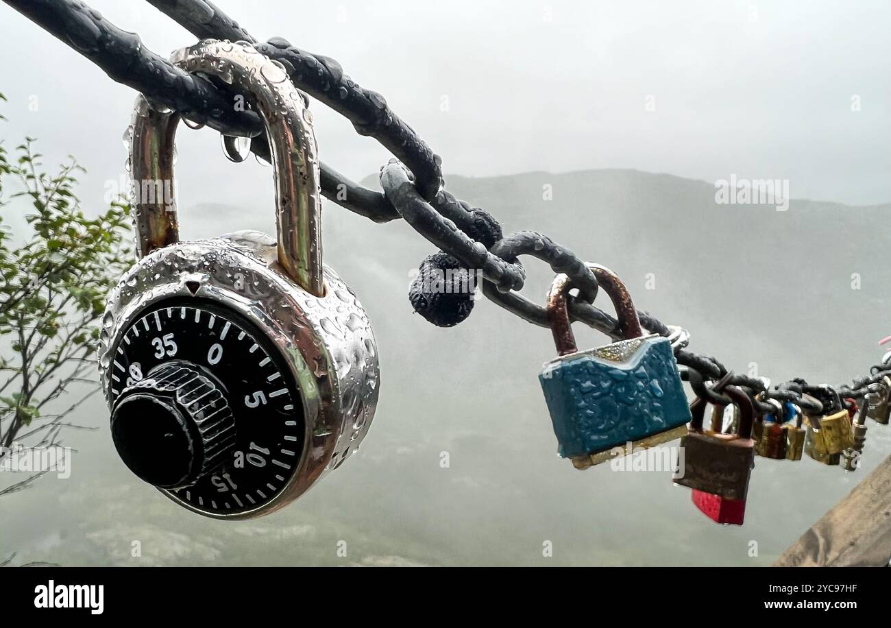 Eine Nahaufnahme mehrerer Vorhängeschlösser und ein Kombinationsschloss, das an einem regnerischen Tag im Freien an einer nassen Kette befestigt ist. Die nebelige, natürliche Landschaft im Hintergrund Stockfoto
