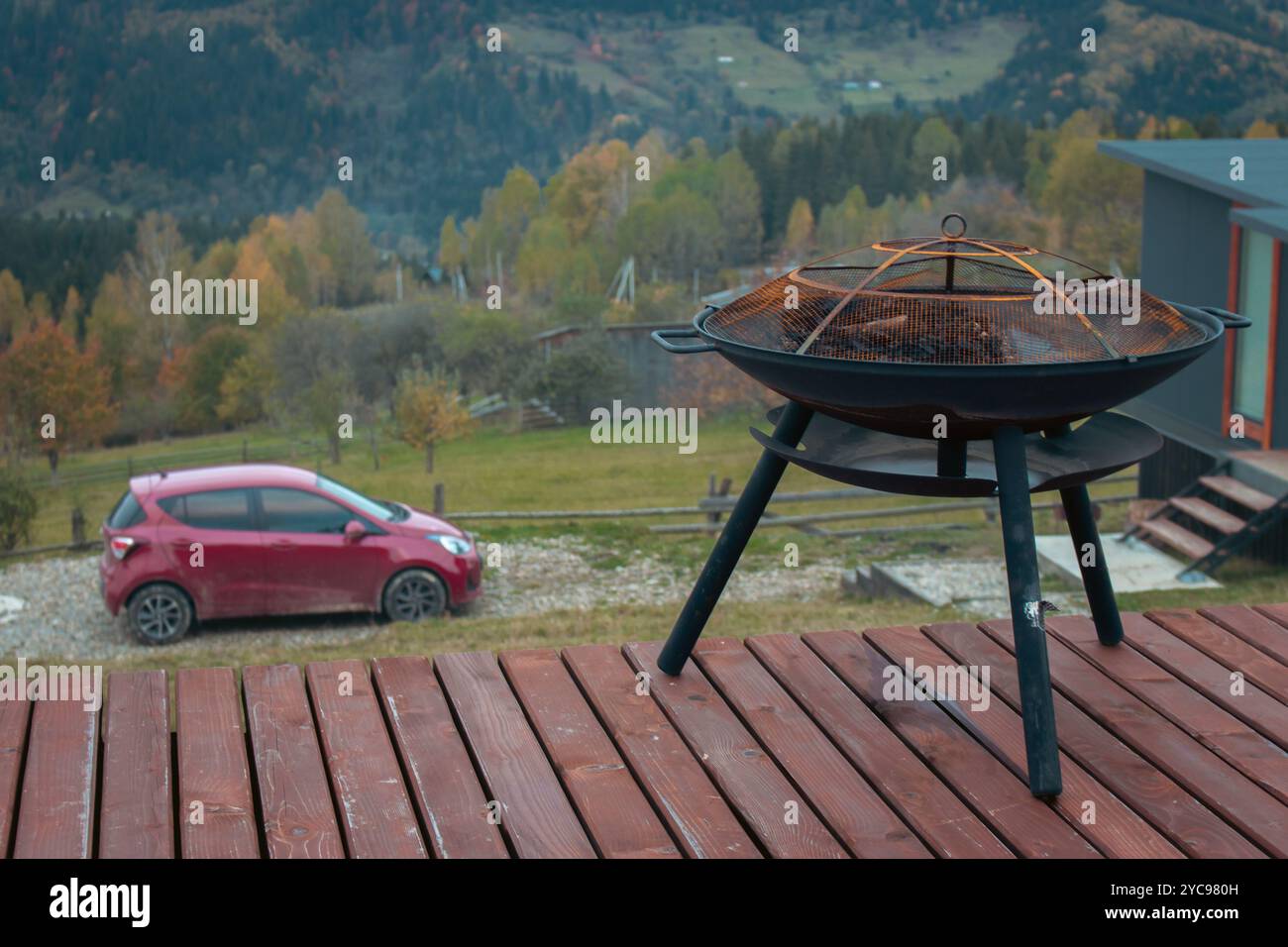 Grillrost auf Holzterrasse mit rotem Auto und Waldhügel im Hintergrund. Grill und Auto auf Schotterstraße in den Bergen. Urlaub in Karpaten Stockfoto