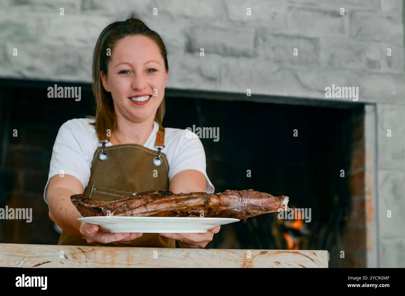 Lächelnde Frau mit einem Lammteller in den Händen, neben einem Grillkoch, mit brauner Schürze und weißem T-Shirt Stockfoto