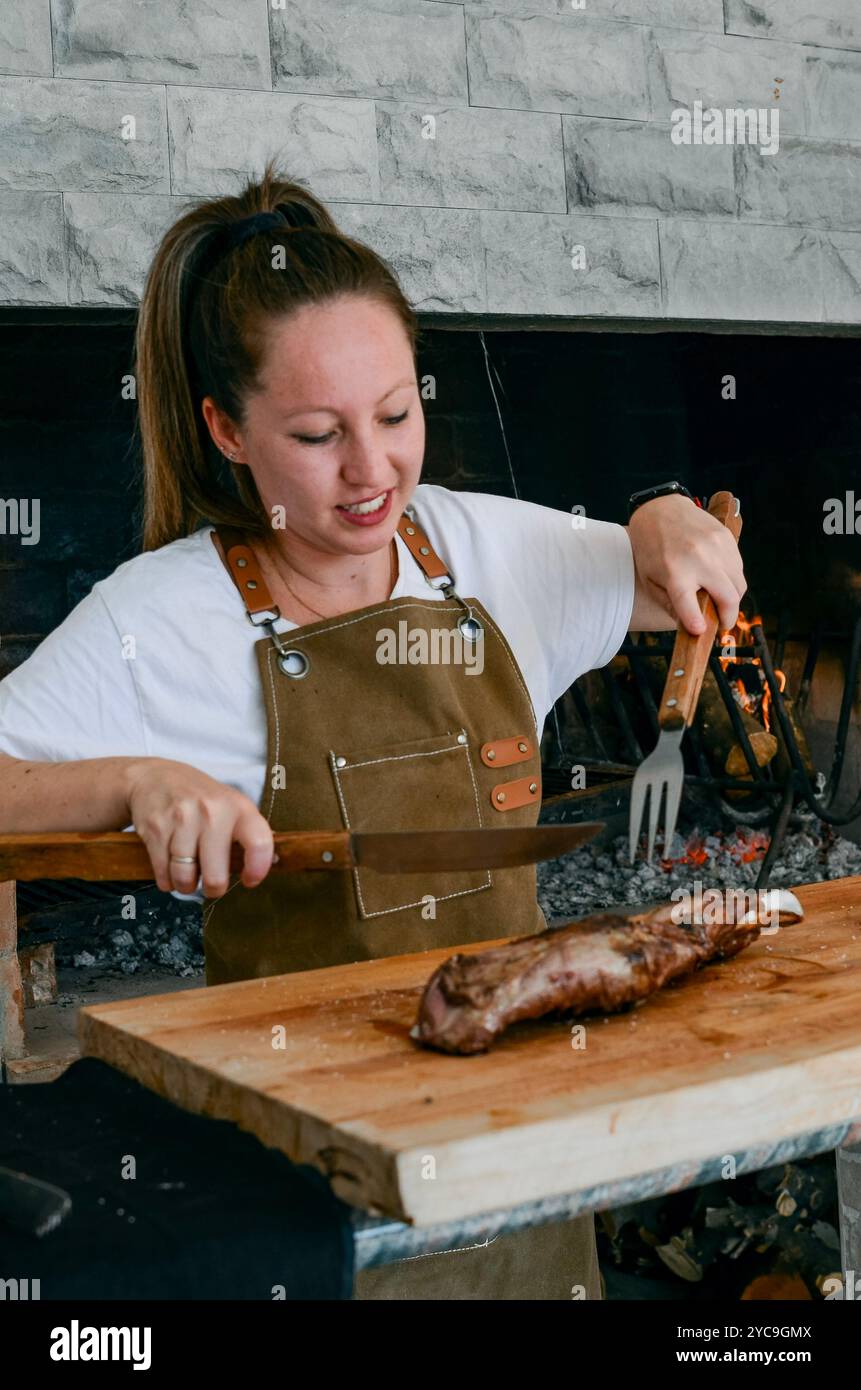 Lächelnde Frau mit hellen Augen, Lammfleisch schneiden, neben einem Grill mit Küchenutensilien, mit brauner Schürze und weißem T-Shirt Stockfoto