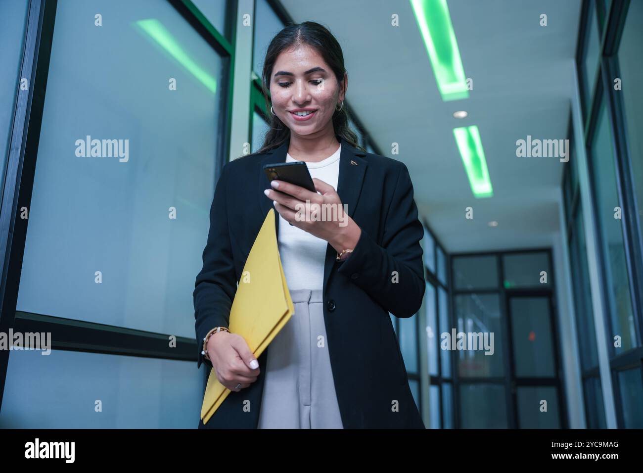 Selbstbewusste junge indische Geschäftsfrau, die durch den Flur des Büros für Meetings geht, während sie mobil ist und Berichte oder Dokumente in der Hand hält. busin Stockfoto