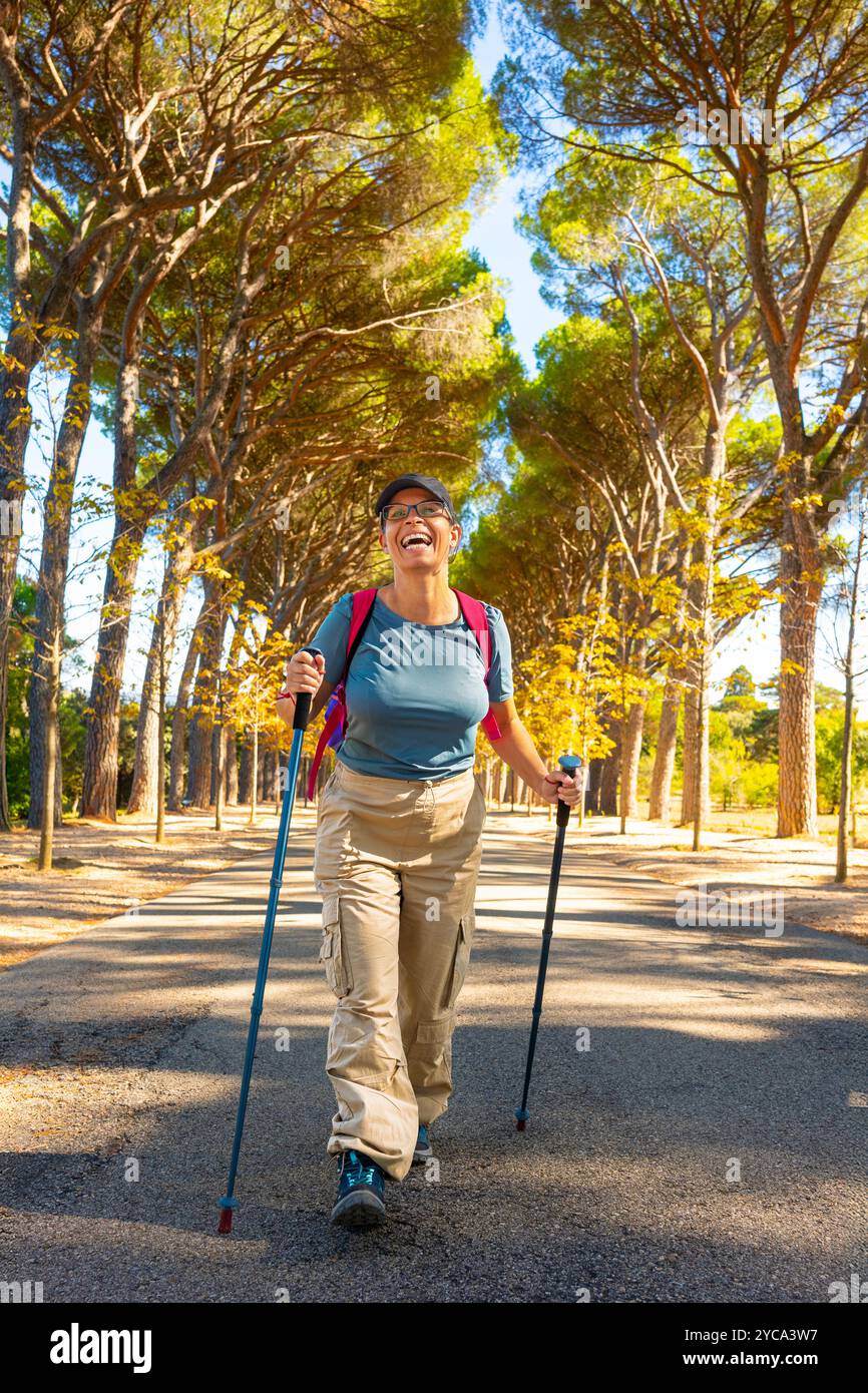 Eine Frau wandert auf einem sonnendurchfluteten Pfad, der von hohen Bäumen gesäumt ist, mit Trekkingstöcken, Trekkingkleidung und Rucksack. Stockfoto