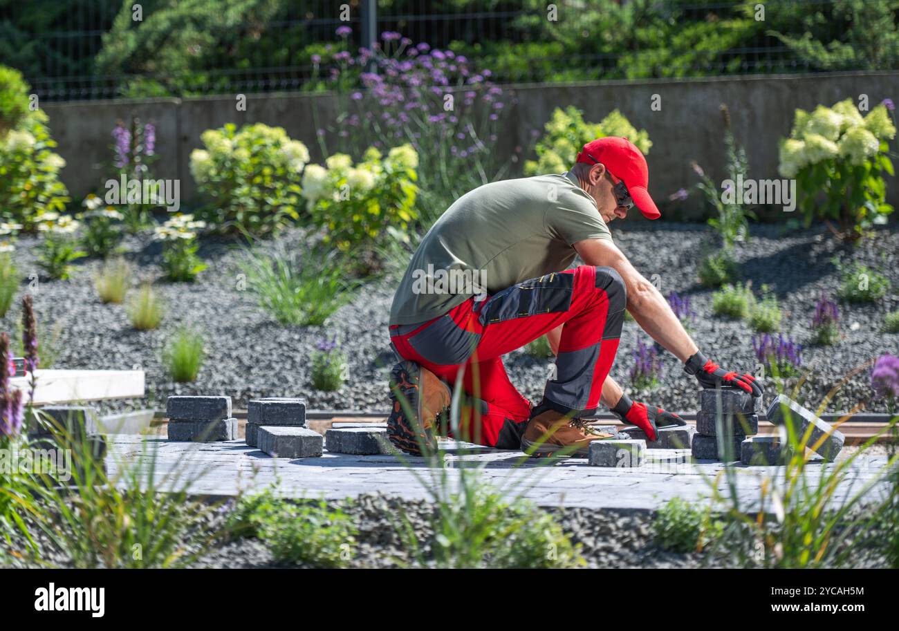 Ein Gärtner arrangiert sorgfältig graue Pflastersteine in einem wunderschön angelegten Garten, umgeben von Grün und blühenden Blumen unter hellen s Stockfoto