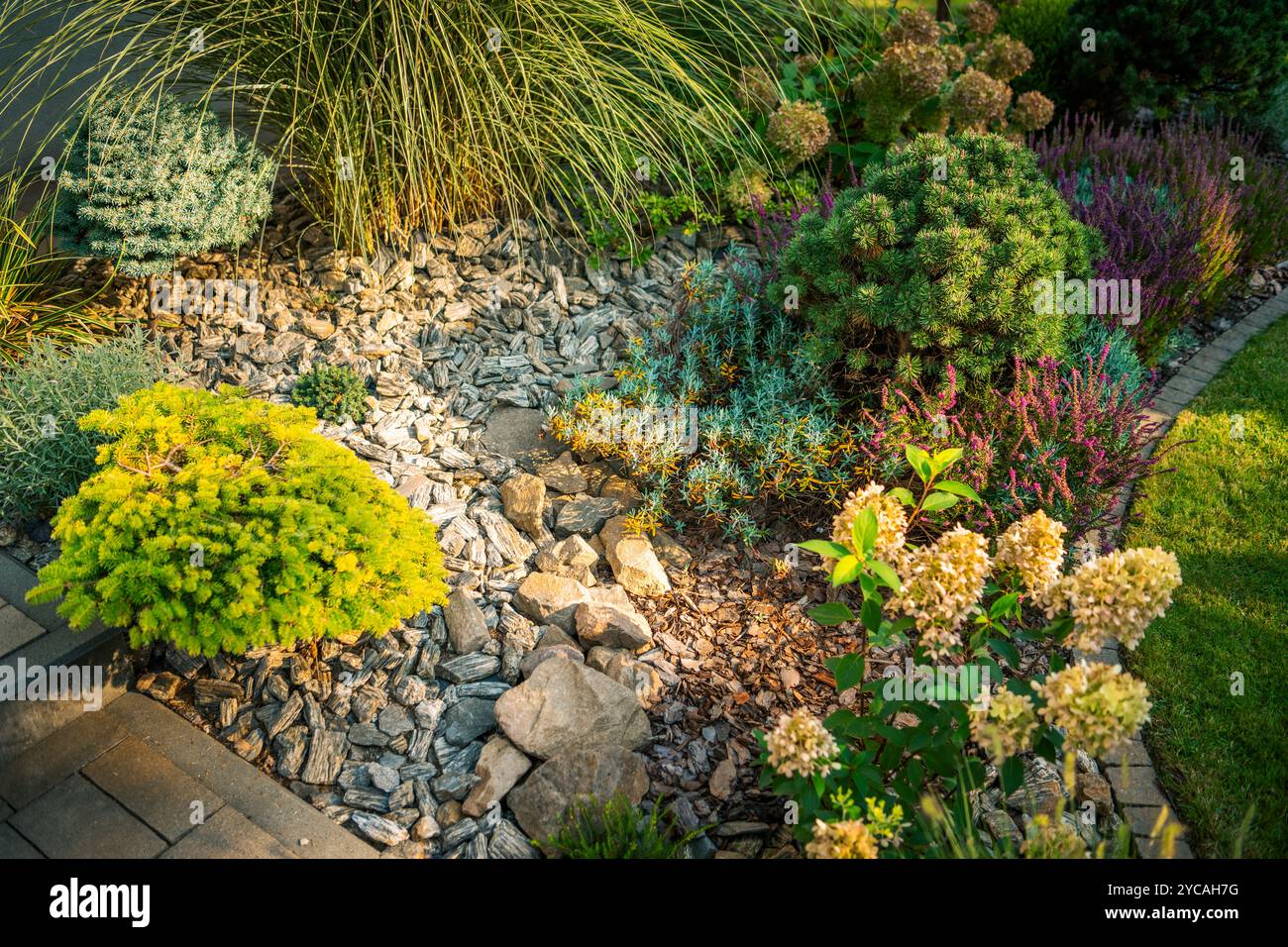 Ein lebendiger Garten bietet eine Mischung aus Grün und blühenden Pflanzen, die von felsigen Wegen unterstrichen und durch das warme Leuchten der frühen Abendsonne verstärkt werden. Stockfoto