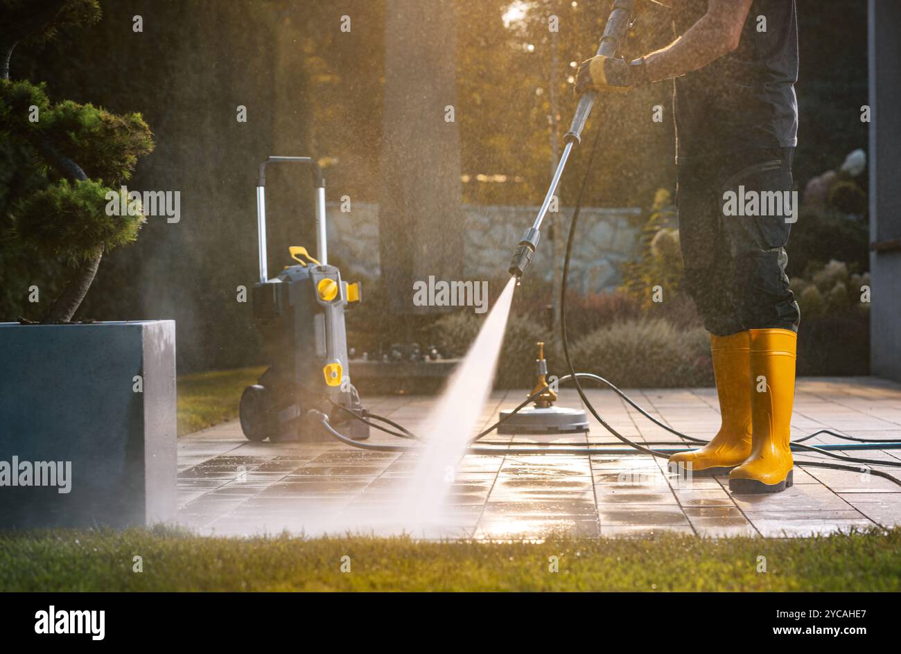 Ein Arbeiter in gelben Stiefeln verwendet einen Hochdruckreiniger, um einen Terrassenbereich unter hellem Sonnenlicht zu reinigen und so eine effektive Wartung im Freien zu gewährleisten. Stockfoto