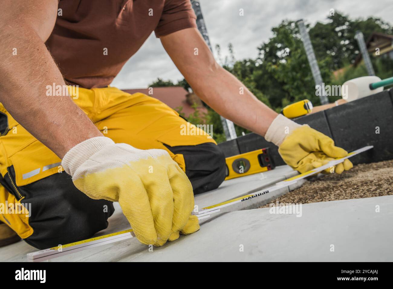 Ein Arbeiter in Handschuhen misst Baumaterialien mit einem Maßband, während er auf dem Boden kniet. Die Einstellung umfasst Werkzeuge, Kies und nahe gelegene g Stockfoto