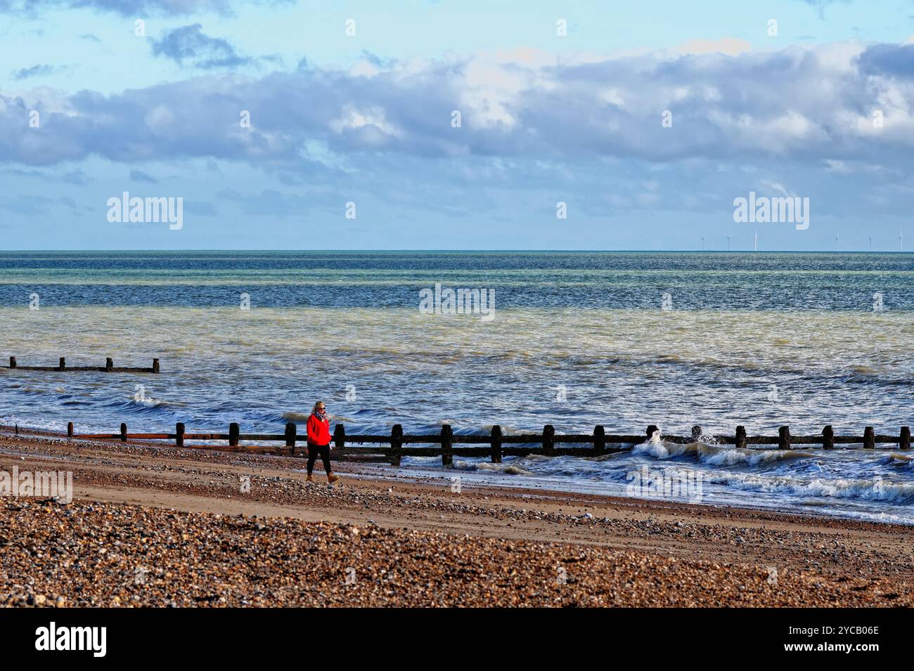Eine einsame Frau in rotem Mantel, die an einem sonnigen Herbsttag am Strand von Littlehampton entlang läuft Stockfoto